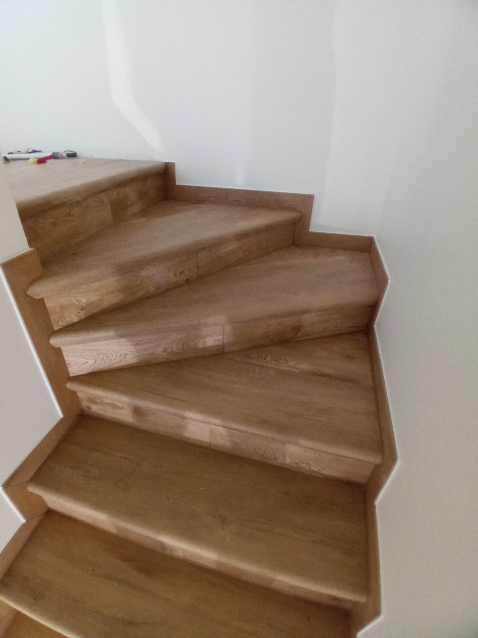 Drewniane schody z wykończeniem w kolorze jasnego dębu, widoczne z góry, z białymi ścianami w tle, częściowo wykończone.