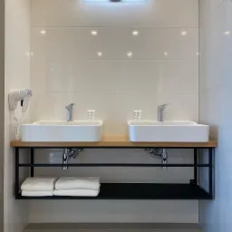 Dwie białe, prostokątne umywalki nablatowe z chromowanymi bateriami, ustawione na blacie z jasnego drewna i czarnym metalowym stelażu. Na dolnej półce stelaża ułożone złożone białe ręczniki.