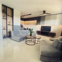 Nowoczesny salon z szarą sofą modułową, stolikami kawowymi z marmurowym blatem, zabudową regałową z drewnianymi półkami i telewizorem na tle zabudowy kuchennej z oświetleniem LED.