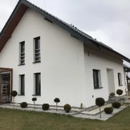 Biały dom jednorodzinny z szarą dachówką i ciemnymi oknami po ociepleniu ścian, z widocznymi elementami rusztowania w tle i małymi drzewkami ozdobnymi wzdłuż ścieżki.