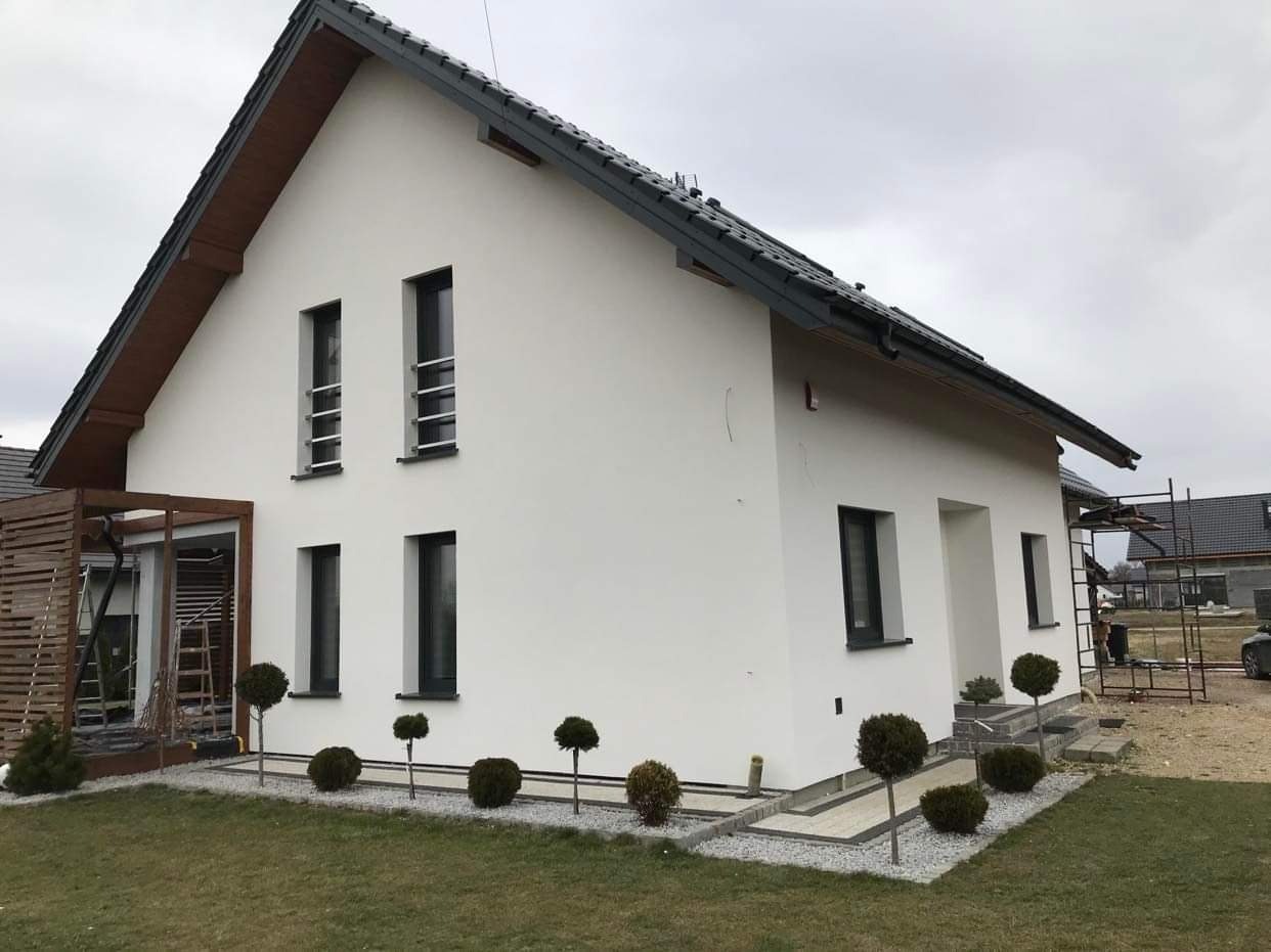 Biały dom jednorodzinny z szarą dachówką i ciemnymi oknami po ociepleniu ścian, z widocznymi elementami rusztowania w tle i małymi drzewkami ozdobnymi wzdłuż ścieżki.