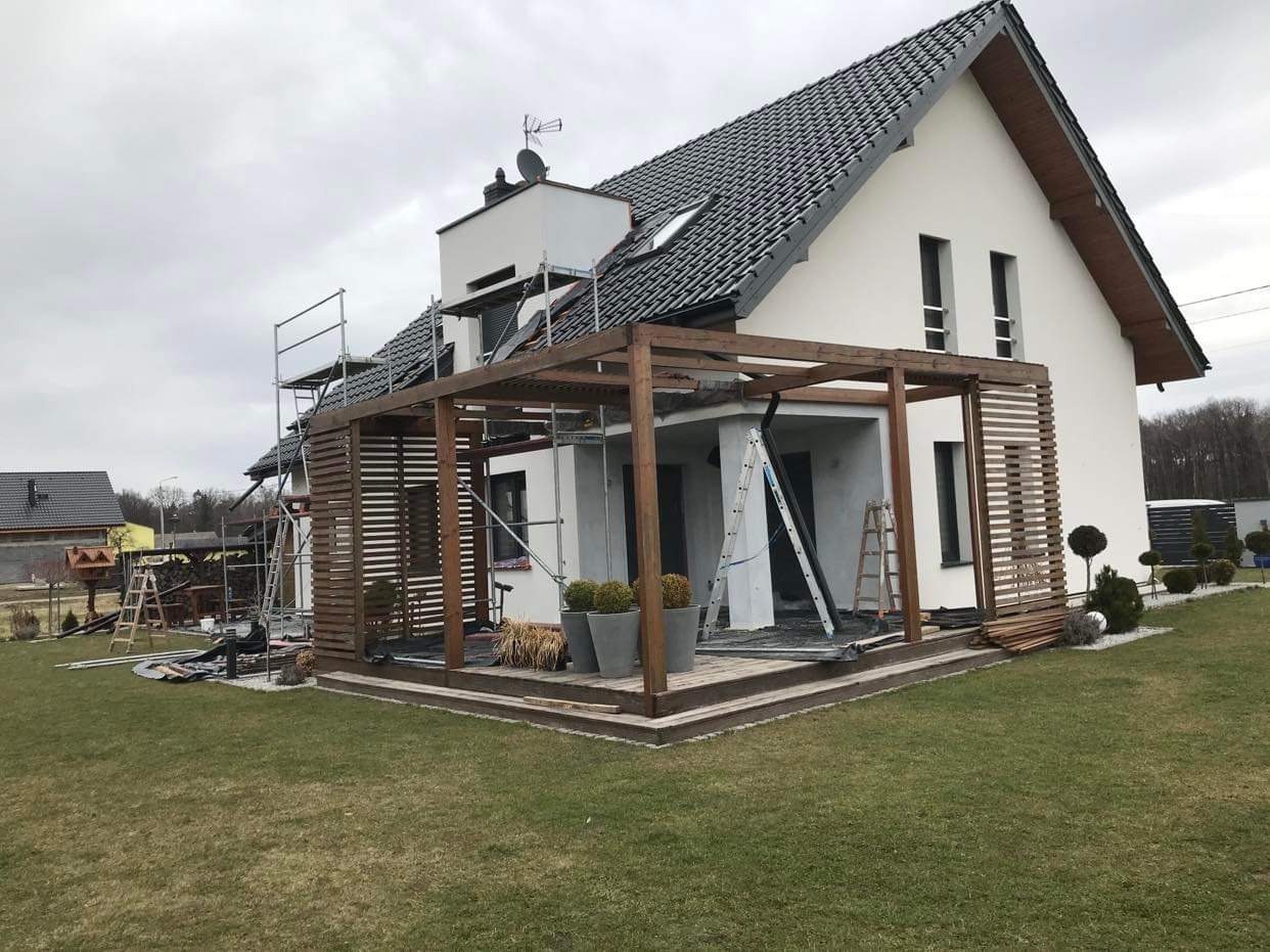 Dom jednorodzinny z drewnianą pergolą w trakcie budowy, widoczne rusztowania i drabiny, trawnik z przodu.