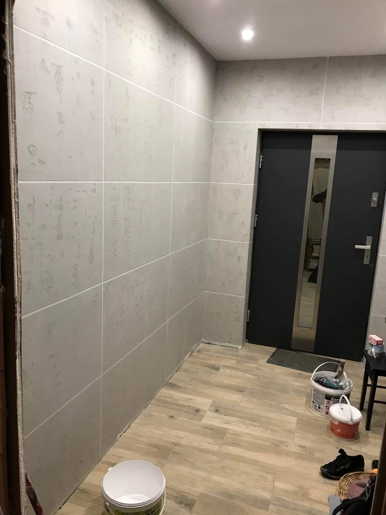 Ściany wykończone szpachlą dekoracyjną w odcieniach szarości, ułożone w kwadraty, z widocznymi wiaderkami farby i narzędziami malarskimi na podłodze imitującej drewno, w pobliżu czarnych drzwi...