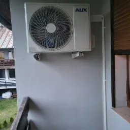 Biała jednostka zewnętrzna klimatyzacji marki AUX zamontowana na szarej ścianie balkonu z widokiem na sąsiedni budynek.