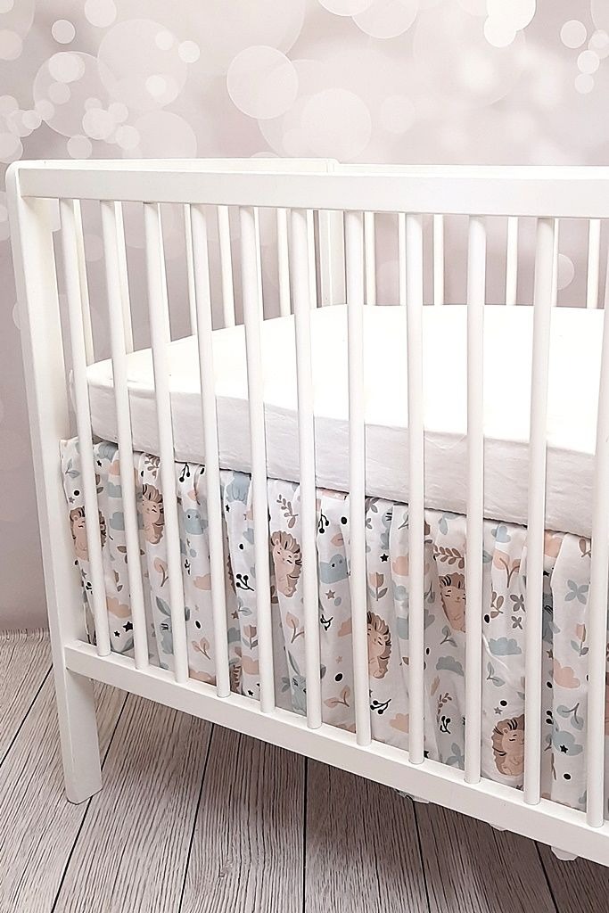 Białe łóżeczko dziecięce z pościelą w pastelowe lwy, chmurki i rośliny, widok z boku na tle ściany w kropki.