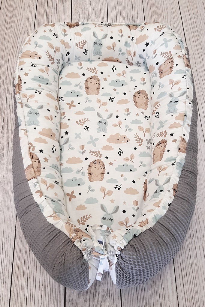 Dziecięce gniazdko niemowlęce z wzorem w króliki, jeże, chmurki i rośliny na białym tle, otoczone szarym, teksturowanym materiałem wafelkowym.