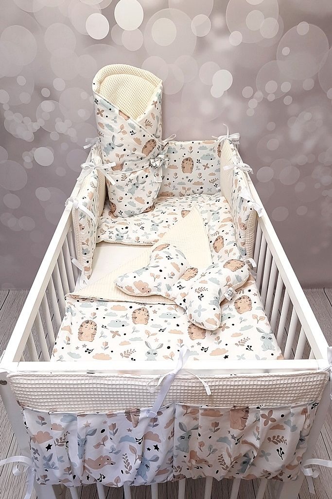 Komplet pościeli dziecięcej w łóżeczku, wzór w jeże, króliczki i chmurki, pastelowe kolory, ochraniacze z falbanką, kołderka, poduszki motylki, tło z efektem bokeh.