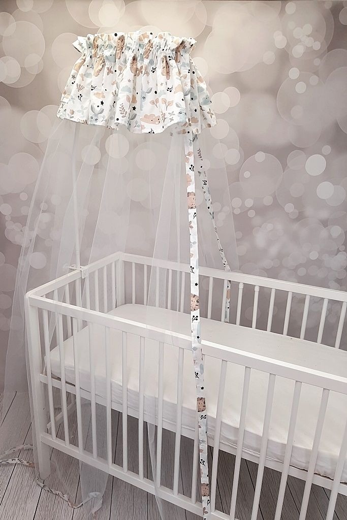 Białe łóżeczko dziecięce z baldachimem z białej tkaniny w pastelowe wzory zwierząt i roślin, na tle ściany w rozmyte kółka.