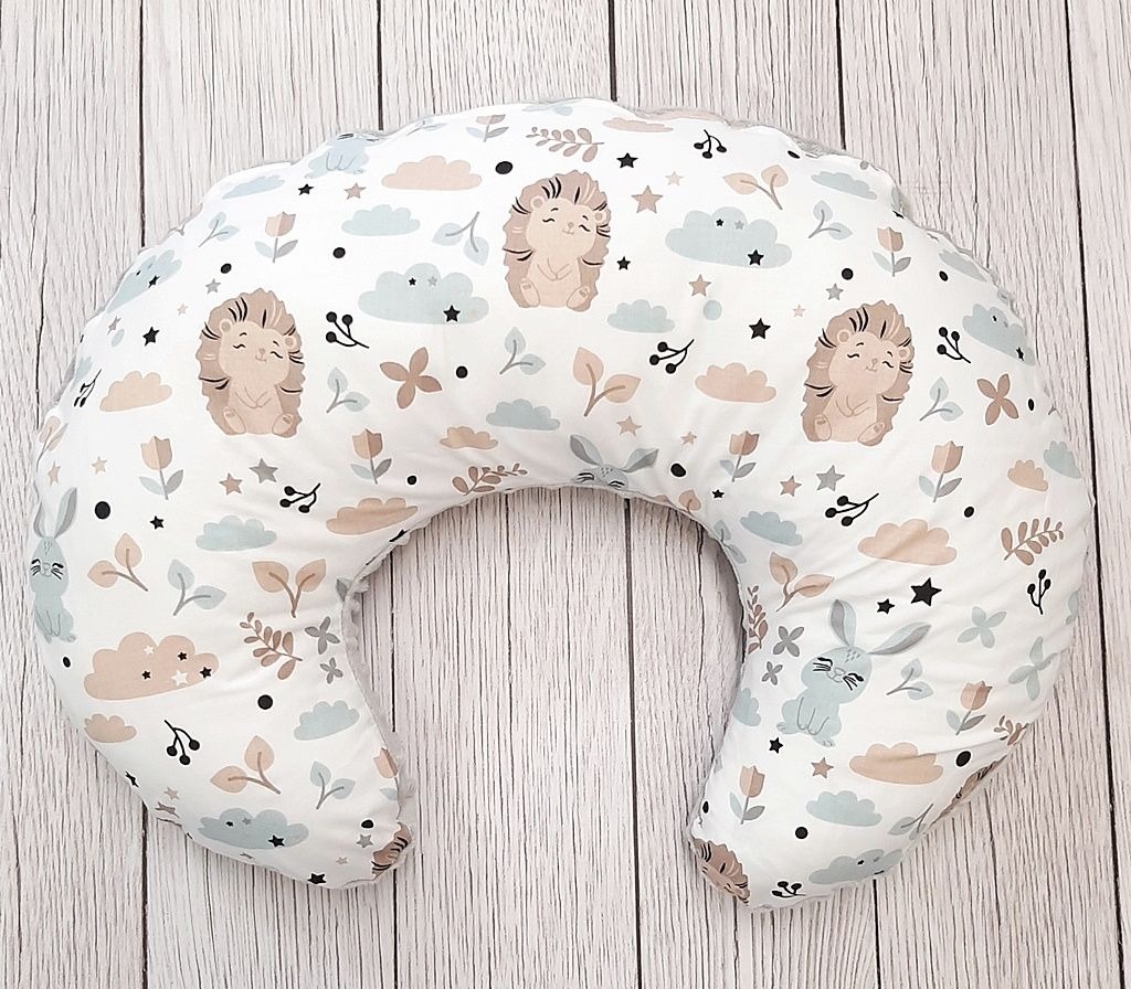 Poduszka do karmienia w kształcie rogala, pokryta białym materiałem w pastelowe wzory: jeże, króliczki, chmurki, kwiatki i gwiazdki, leżąca na drewnianym podłożu.