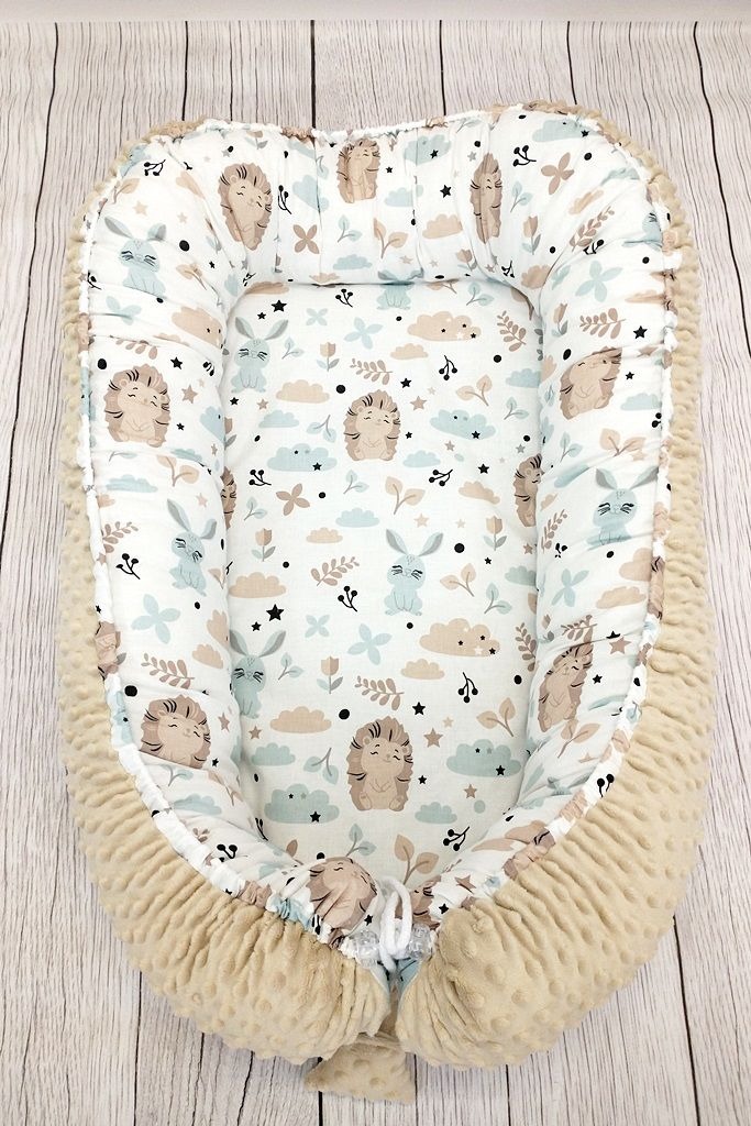 Beżowy kokon niemowlęcy z wzorem w jeże, króliki, chmurki i gwiazdki na drewnianym tle.