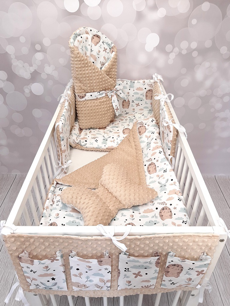 Komplet pościeli dziecięcej z motywem jeży, wypełniający białe łóżeczko, z ochraniaczem z kieszeniami, rożkiem niemowlęcym i poduszkami z wypustkami, na tle rozmytych światełek.