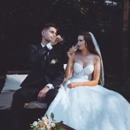 Elegancka para młoda w plenerze pije szampana. Ona w koronkowej sukni z welonem, on w czarnym garniturze. Na stoliku leży bukiet ślubny.