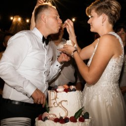 KaluckiStudio Jakub Kałucki - Para młoda karmiąca się kawałkiem dwupiętrowego tortu weselnego z literami 'P&S', owocami i makaronikami, w nocnej scenerii z girlandą światełek tworzących efekt gwiazdy.