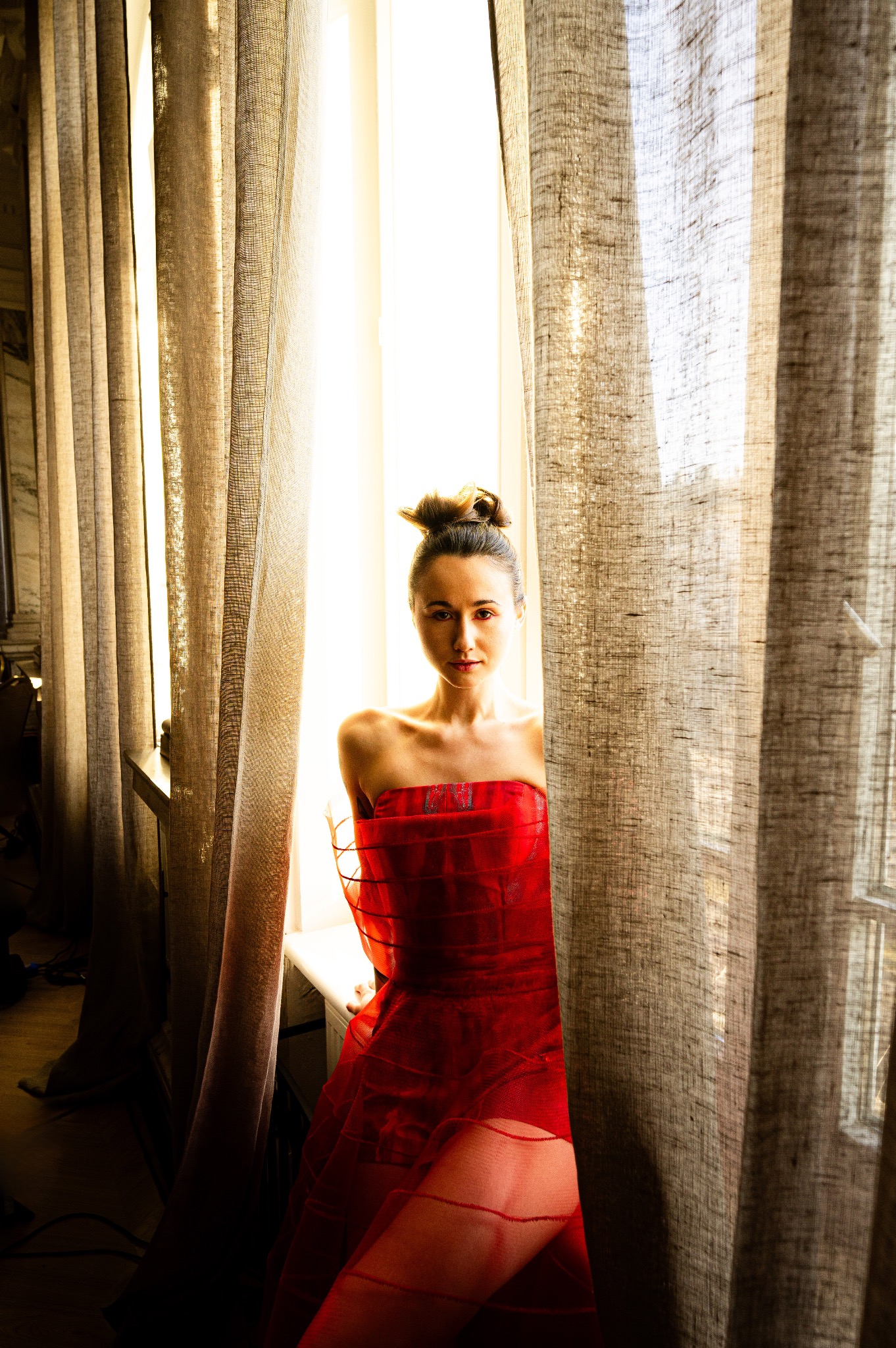 Portret kobiety w czerwonej sukni, stojącej przy oknie z zasłonami. Mocne, naturalne światło podkreśla jej rysy i fakturę materiału. Upięte włosy dodają elegancji.