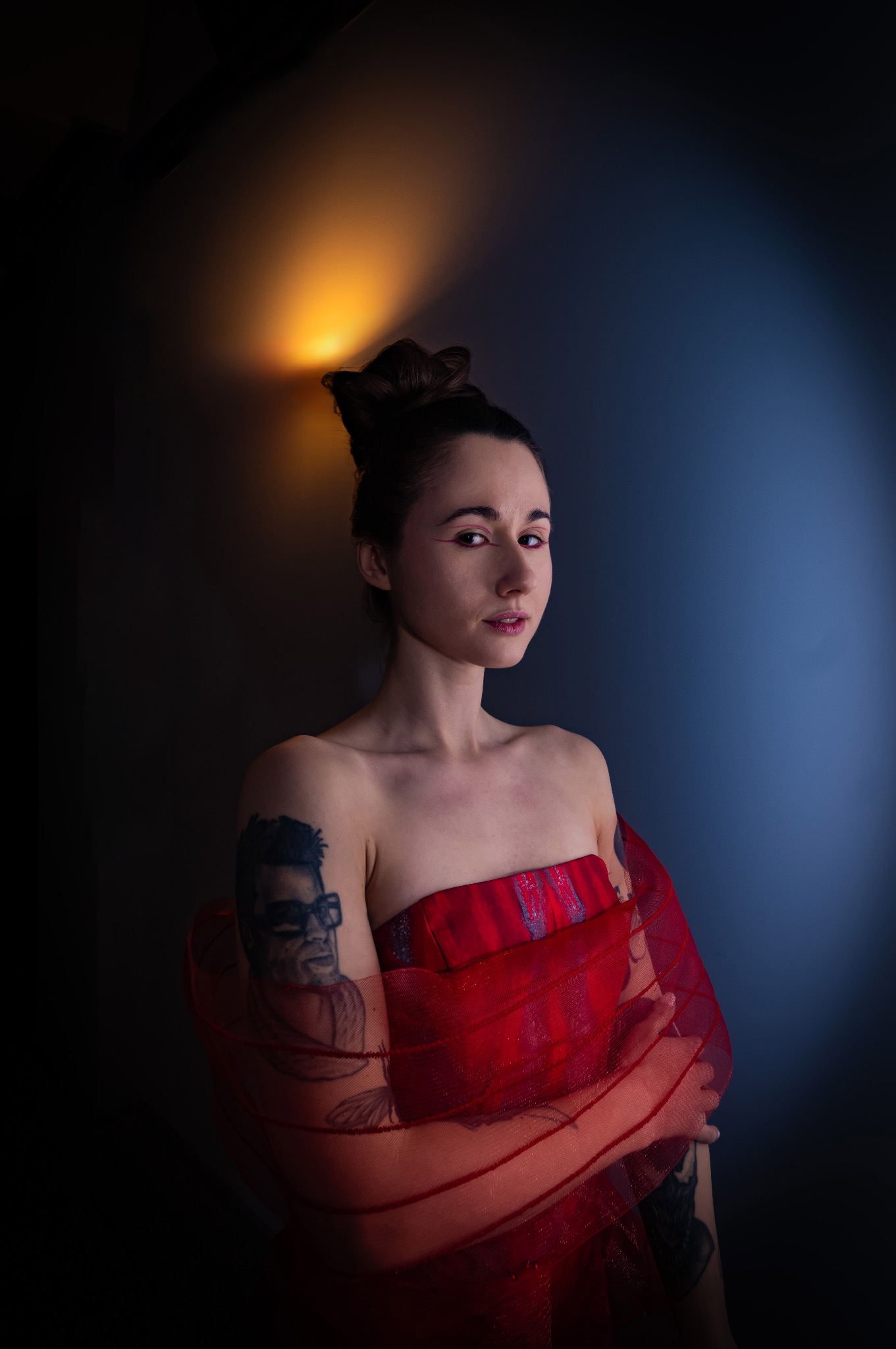 Portret kobiety w czerwonej, przezroczystej tkaninie, z tatuażem na ramieniu, oświetlonej teatralnym światłem na ciemnym tle. Uwagę przykuwa makijaż oczu.
