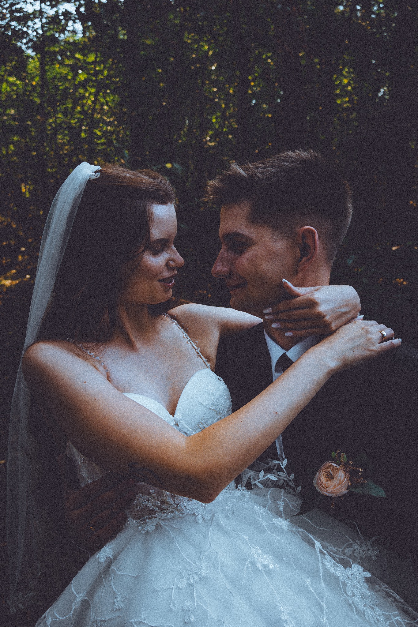 Uśmiechnięta panna młoda w objęciach pana młodego, w tle zieleń drzew. Delikatny welon i kwiecista butonierka dodają uroku.