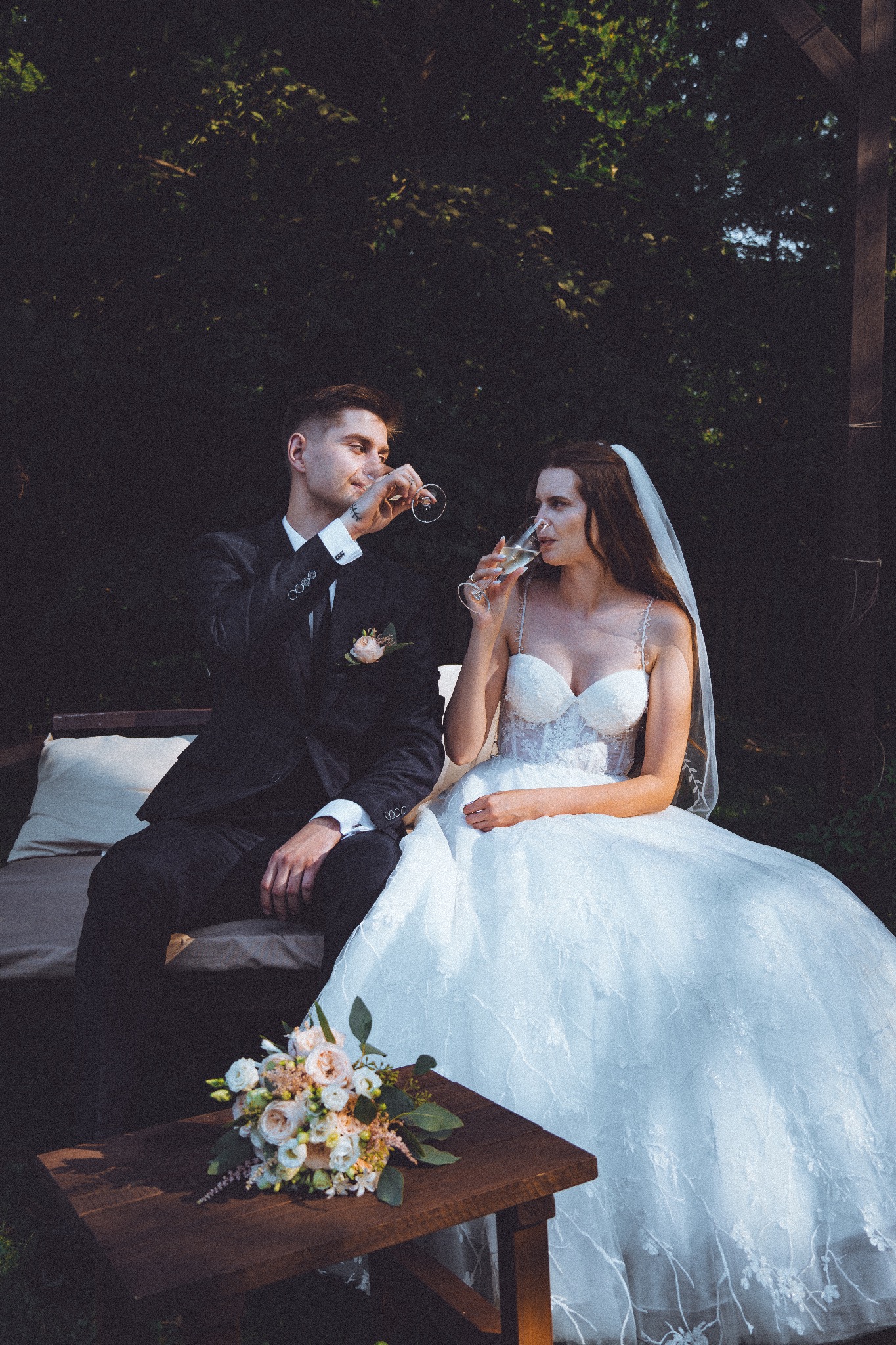 Elegancka para młoda w plenerze pije szampana. Ona w koronkowej sukni z welonem, on w czarnym garniturze. Na stoliku leży bukiet ślubny.