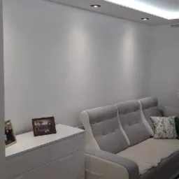 Jasne, świeżo pomalowane wnętrze pokoju w Opolu z podwieszanym sufitem z oświetleniem LED, białą komodą z ramkami i szarą sofą z poduszkami.