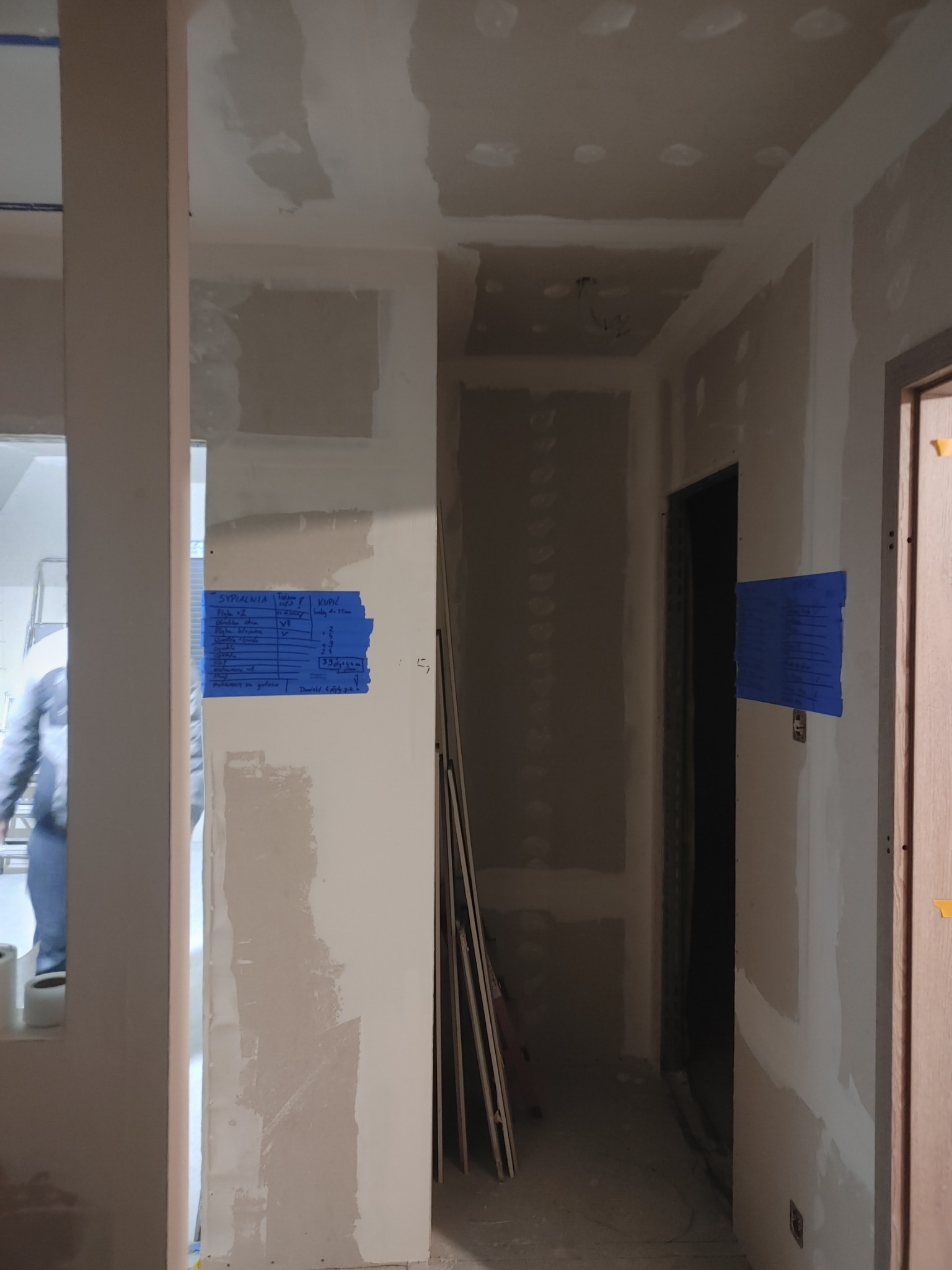 Wnętrze w trakcie prac wykończeniowych, ściany przygotowane do malowania, widoczne szpachlowane powierzchnie i notatki z wymiarami na niebieskiej taśmie malarskiej.