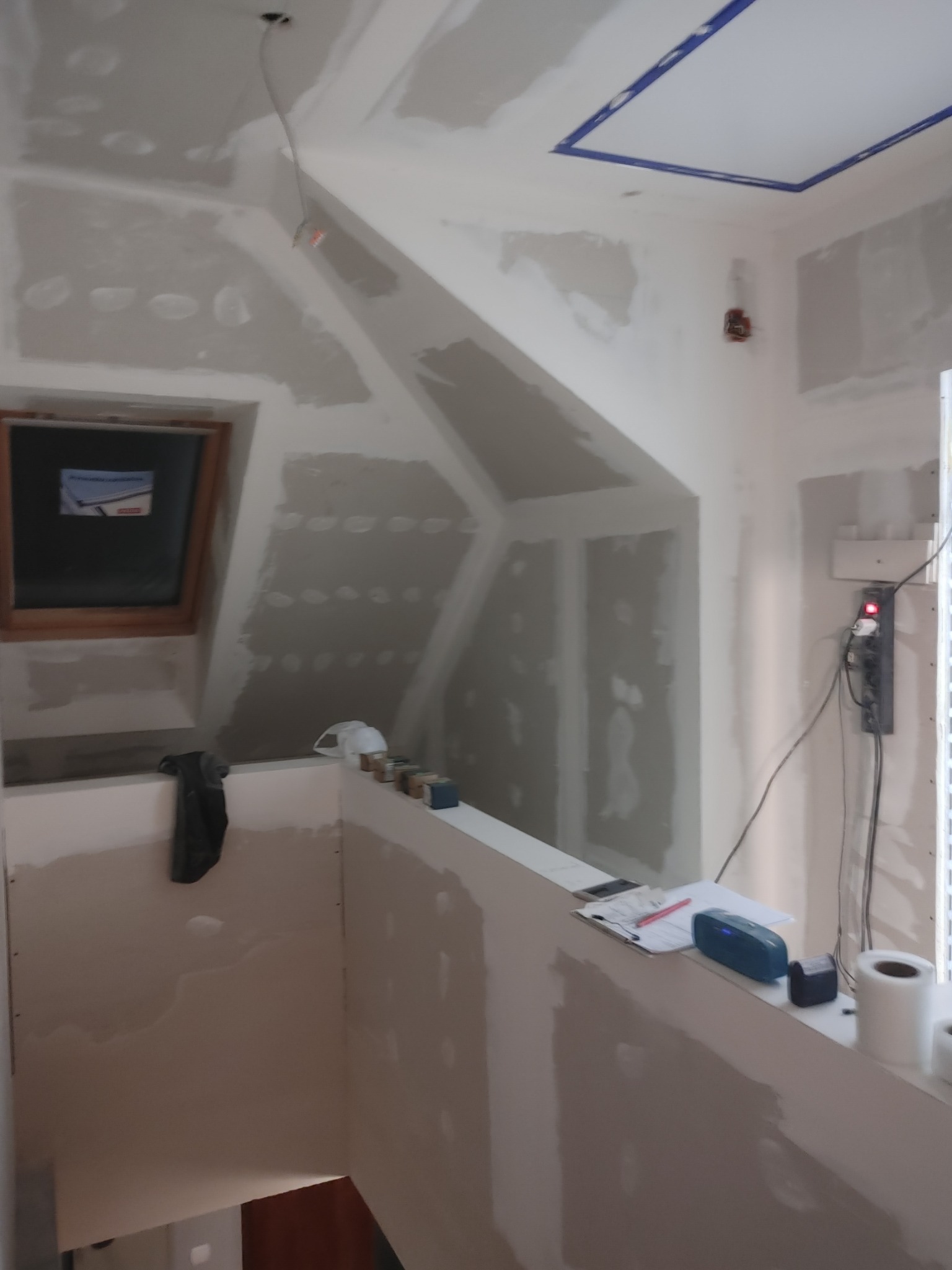 Poddasze w trakcie ocieplania i wykańczania, widoczne płyty karton-gips, okno dachowe, listwa zasilająca, głośnik bluetooth, notatnik z długopisem i rolka taśmy malarskiej.