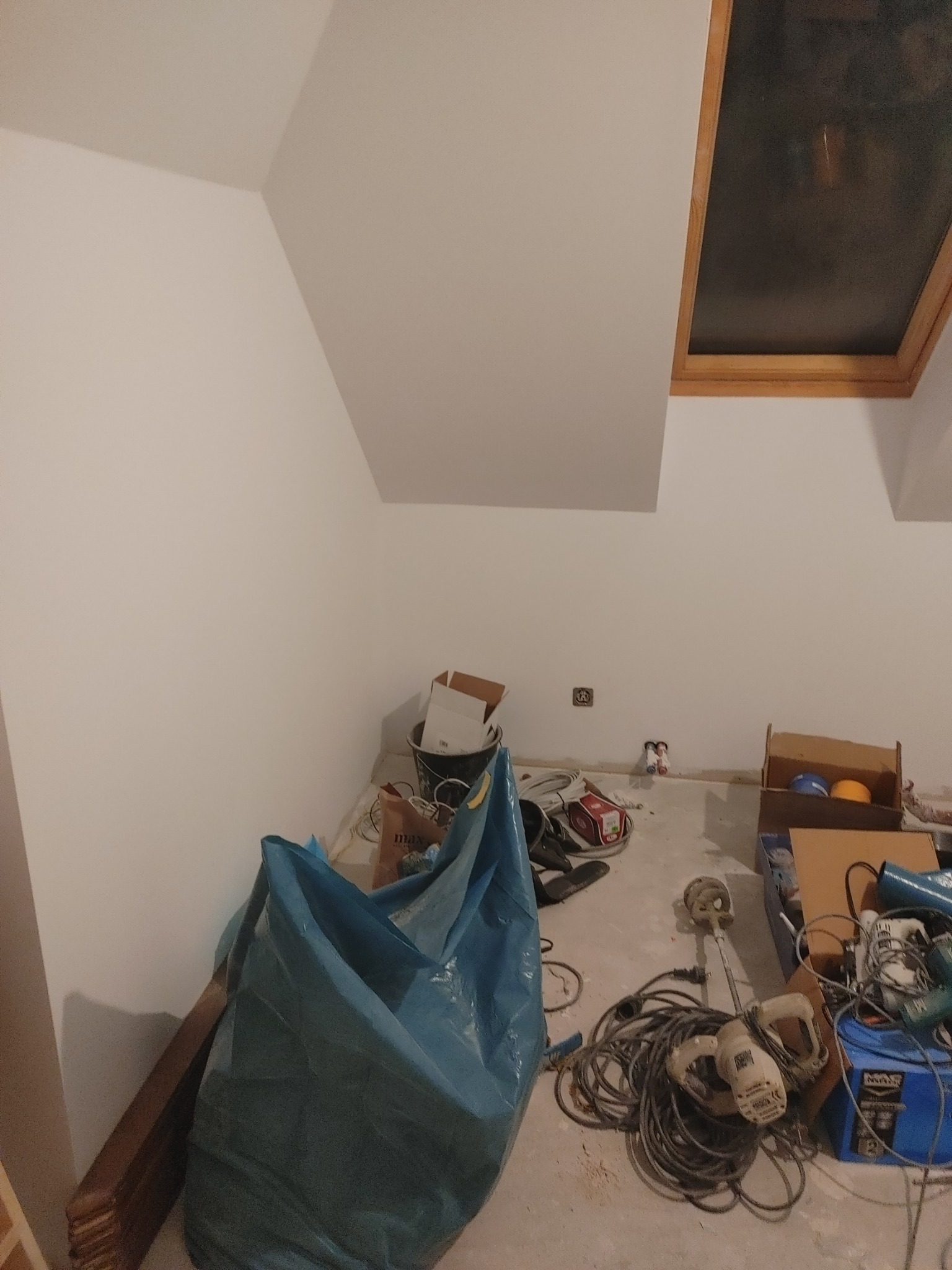 Poddasze w trakcie prac wykończeniowych: białe ściany, okno dachowe, narzędzia budowlane, worki z materiałami, widoczne przewody elektryczne i puszki instalacyjne.