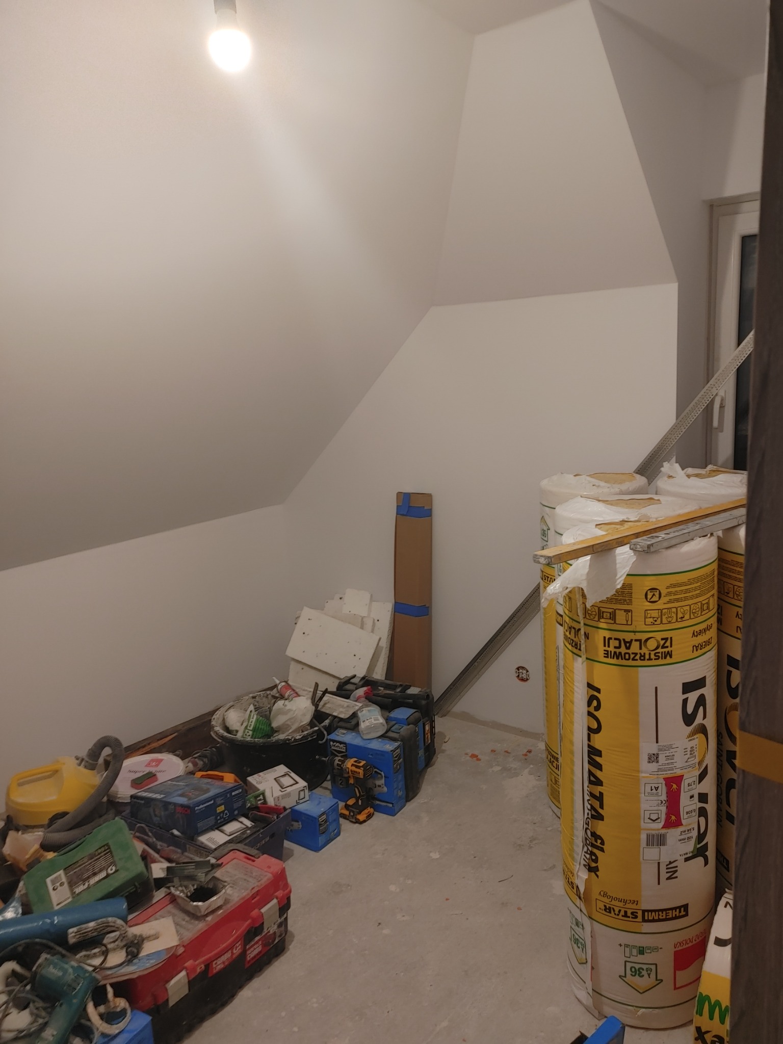 Poddasze w trakcie prac izolacyjnych, widoczne rolki wełny mineralnej Isover, narzędzia i materiały budowlane porozrzucane na podłodze, surowe ściany pod skosem.
