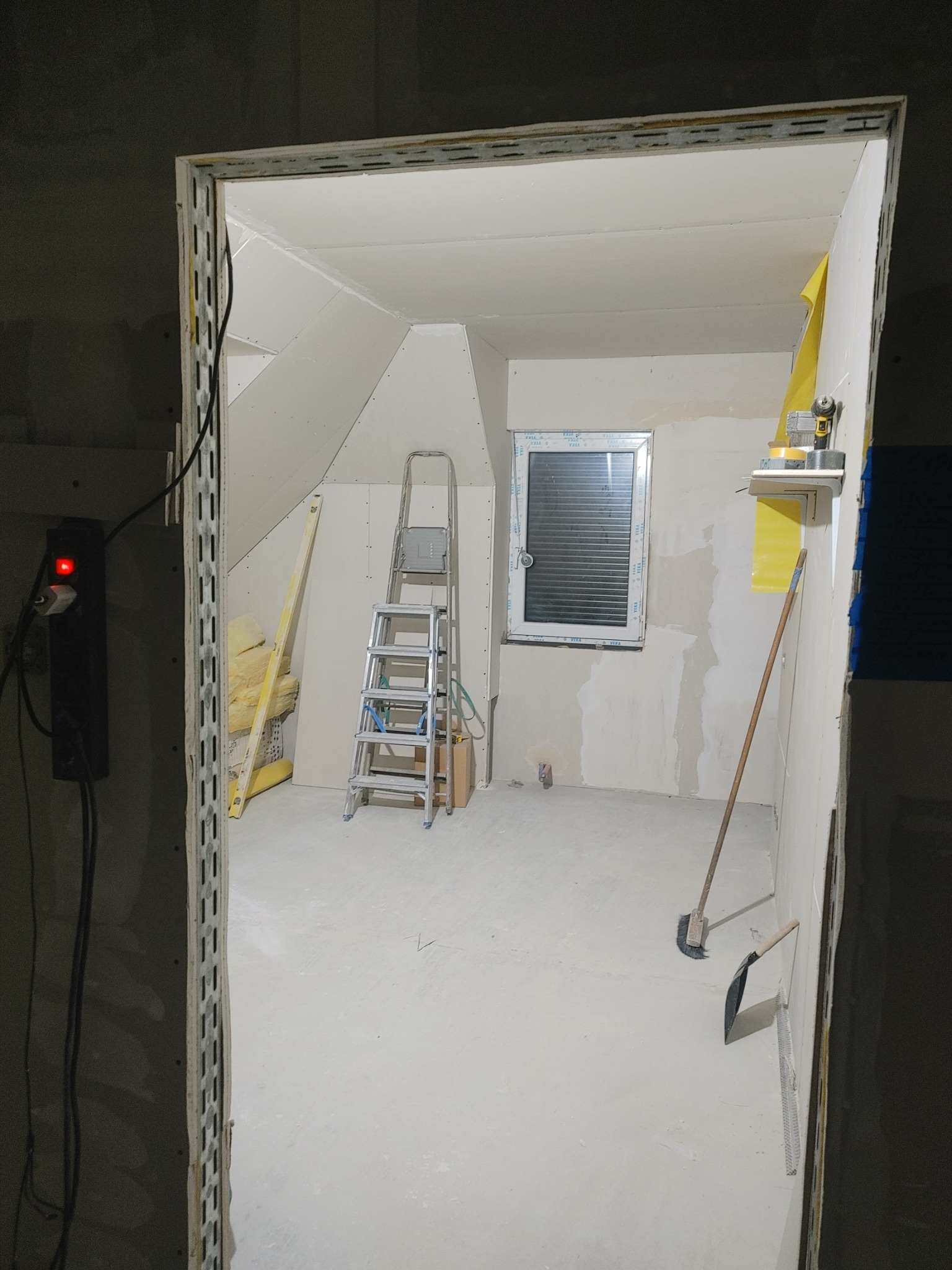 Poddasze w trakcie wykańczania: widoczna drabina, płyty gipsowo-kartonowe na skosach, okno z żaluzjami, poziomica oparta o ścianę, ocieplenie z wełny mineralnej w rogu, oraz narzędzia do sprzątania...