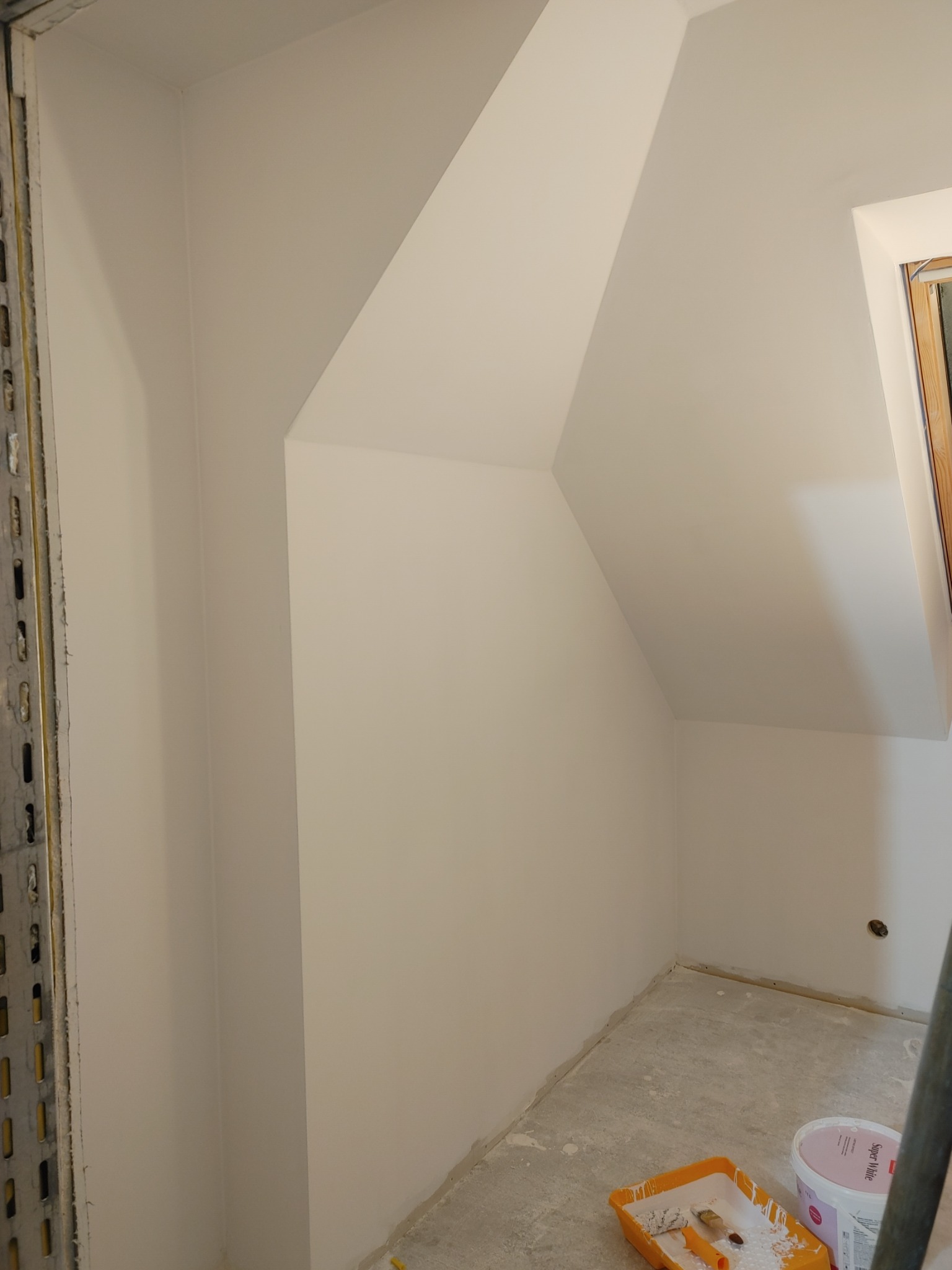 Pomalowane na biało poddasze ze skosem, widoczna kuweta z wałkiem i pędzlem oraz wiadro z farbą Super White, surowa podłoga.