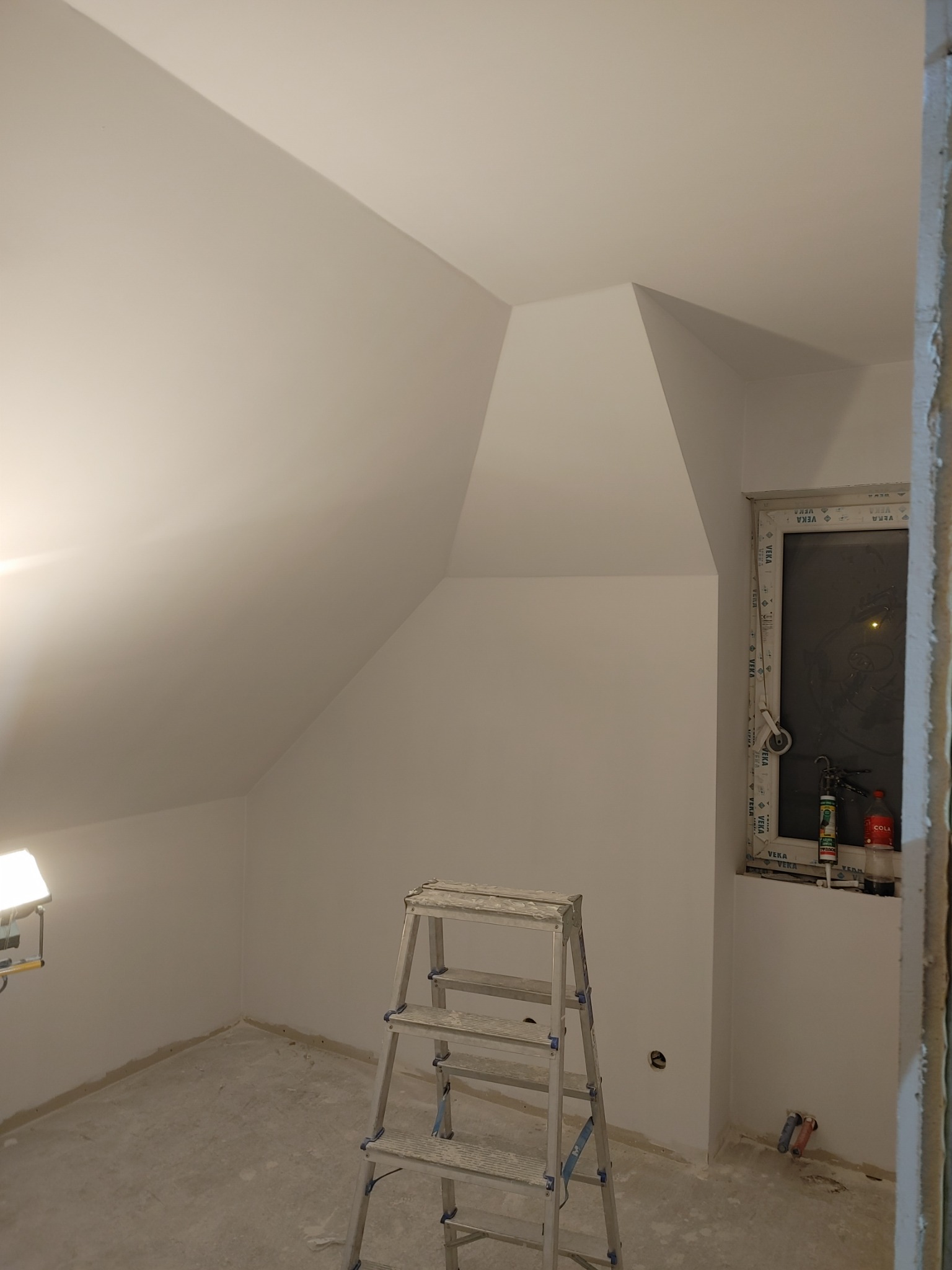 Poddasze po gładzi szpachlowej, przygotowane do dalszych prac wykończeniowych, z drabiną aluminiową na pierwszym planie i oknem PCV z widocznymi uszczelniaczami oraz lampą budowlaną po lewej stronie.