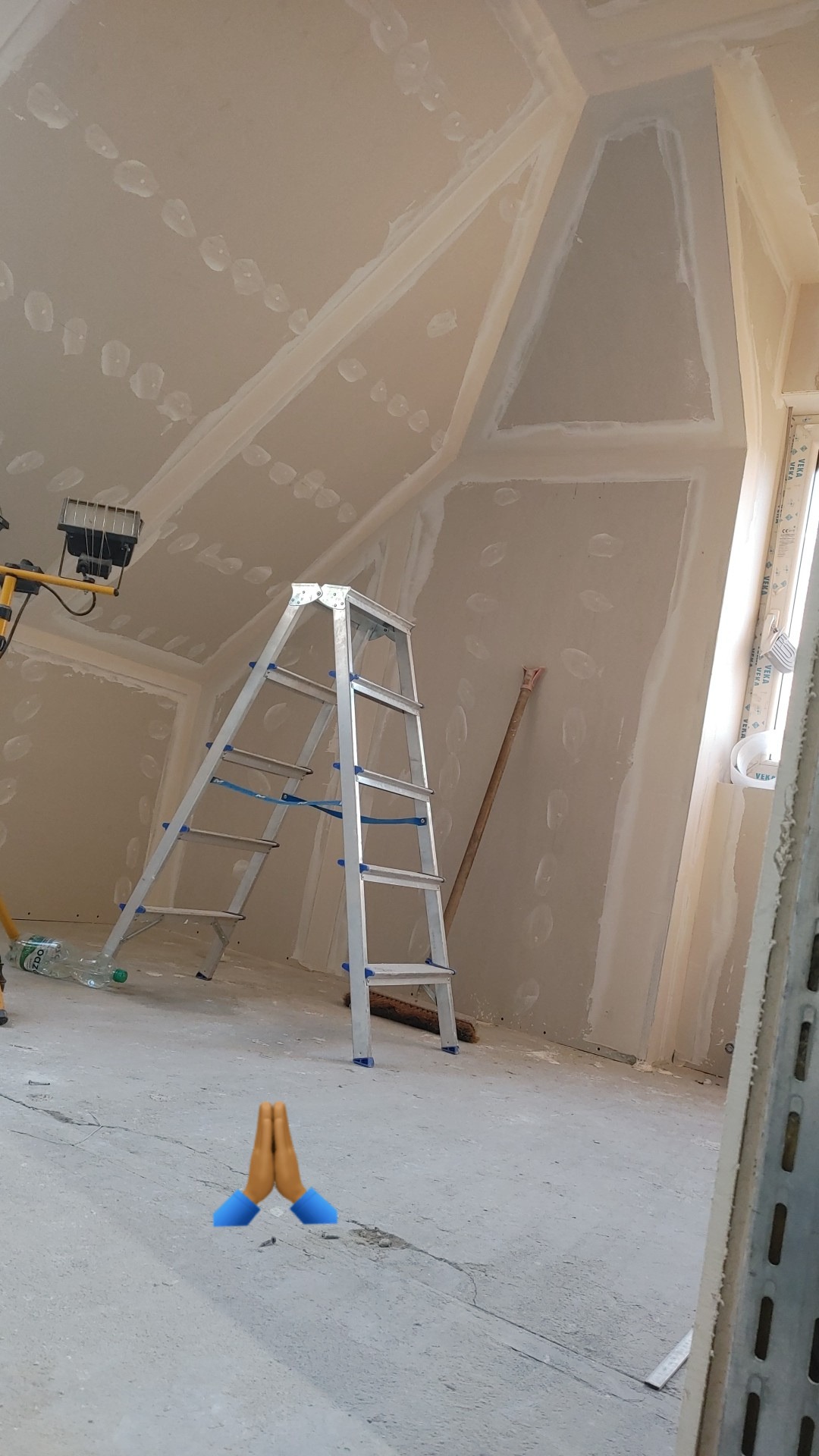 Poddasze w trakcie wykańczania płytami gipsowo-kartonowymi, widoczna aluminiowa drabina, lampa budowlana i ślady po szpachlowaniu na suficie skośnym.