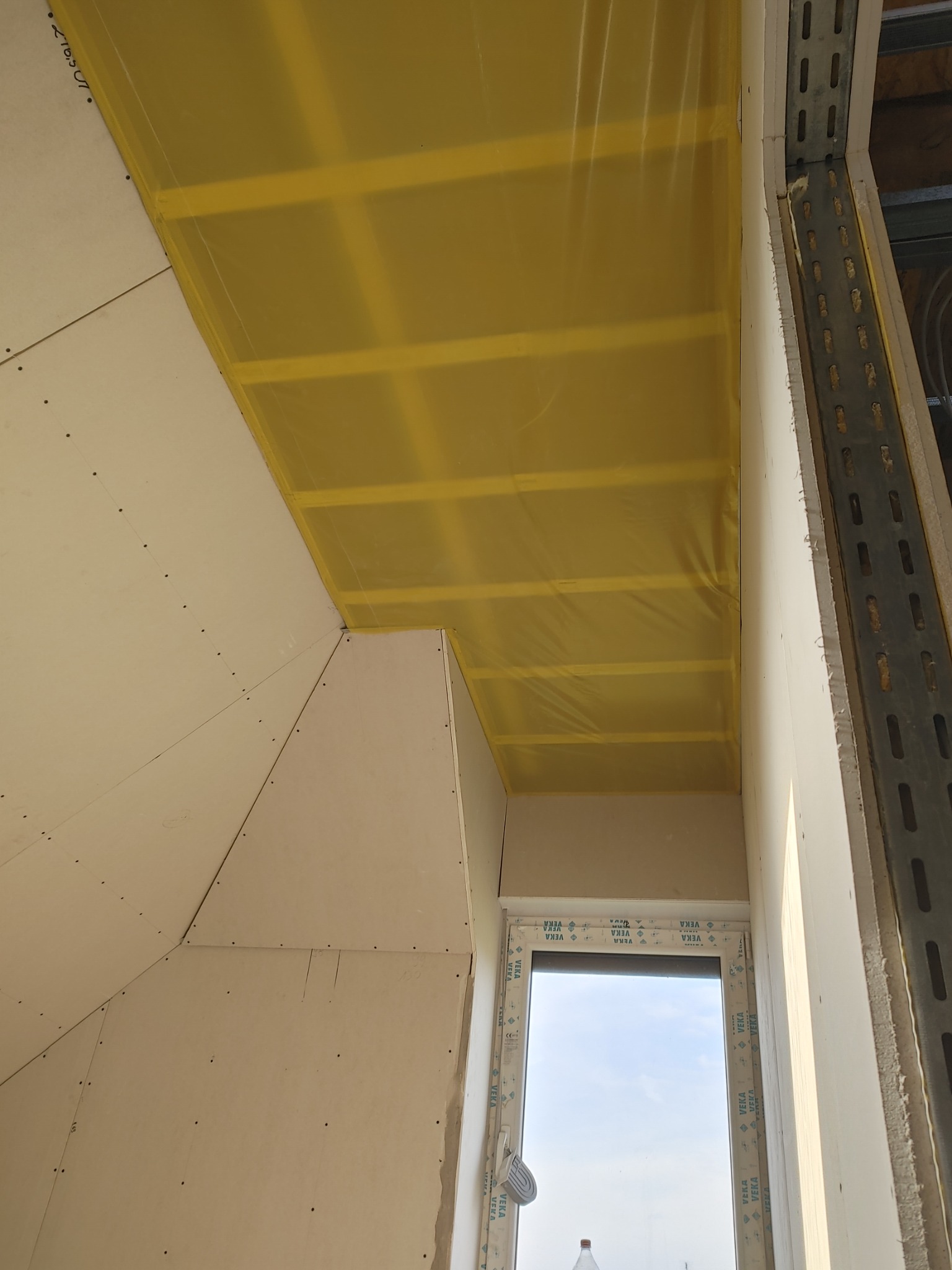 Poddasze w trakcie prac ociepleniowych, widoczne żółte folie paroizolacyjne pod płytami gipsowo-kartonowymi, surowe wykończenie ścian i metalowe profile mocujące wzdłuż okna.