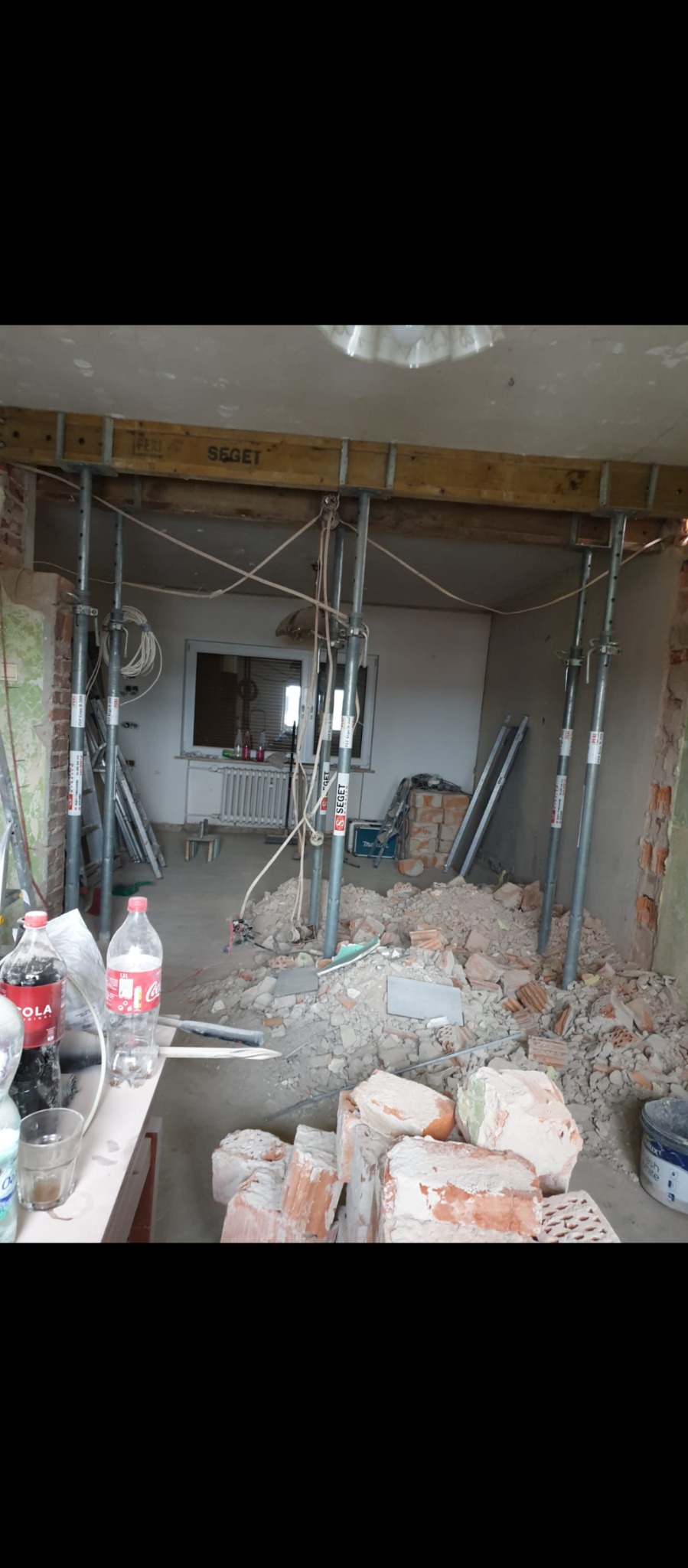 Wnętrze w trakcie przebudowy: widoczne metalowe podpory podtrzymujące drewniane belki stropowe, gruz ceglany na podłodze, butelki z napojami i narzędzia w tle.