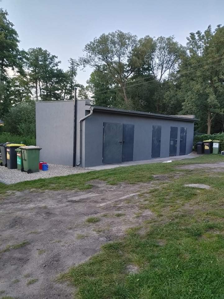 Szary budynek gospodarczy z czterema metalowymi drzwiami, otoczony zielenią, z widocznymi pojemnikami na śmieci w tle.