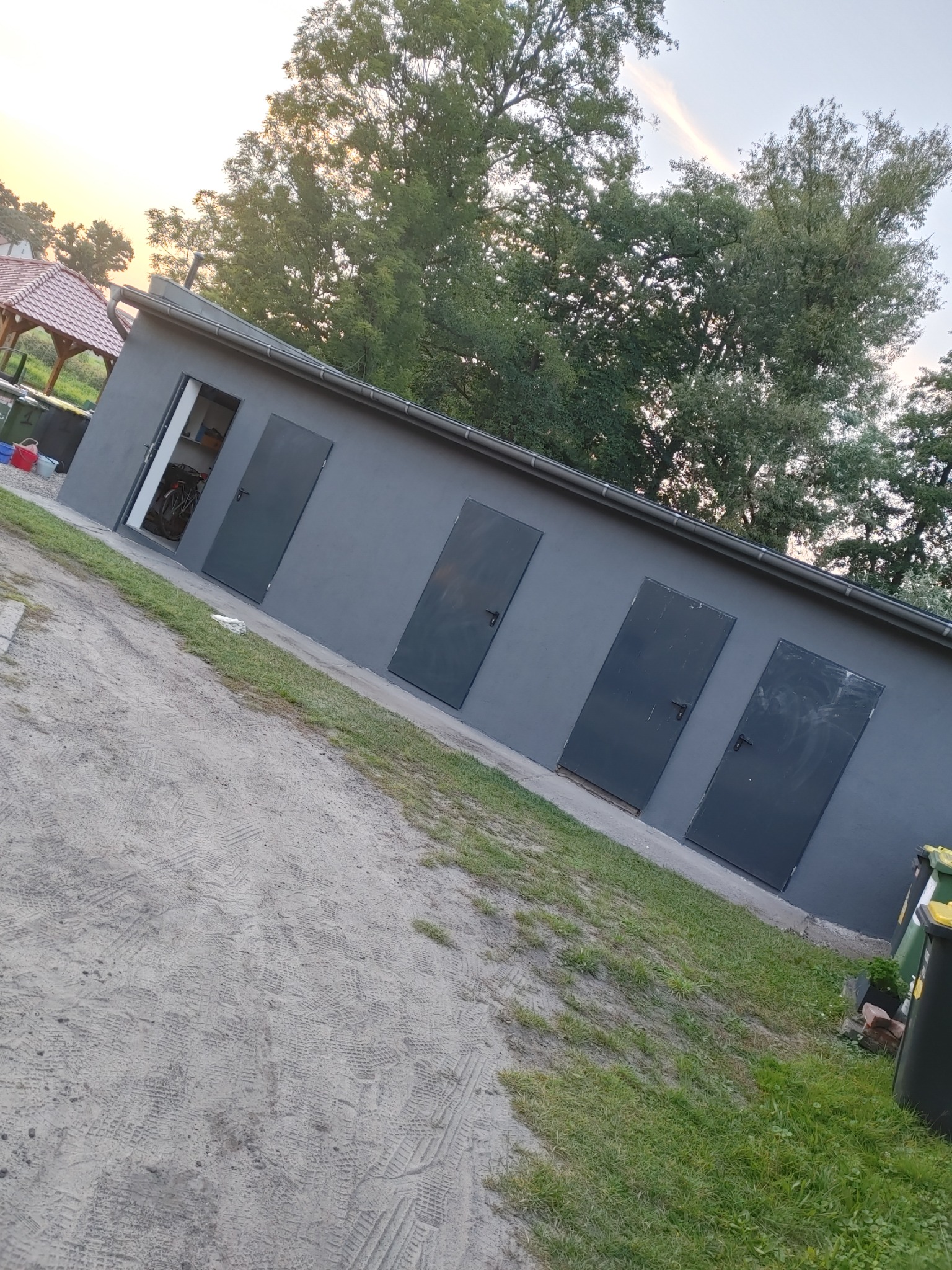 Szary, pomalowany budynek gospodarczy z czterema ciemnymi drzwiami, z których jedne są otwarte, ukazując rower w środku, otoczony piaskową ścieżką i zielenią, sfotografowany pod kątem.