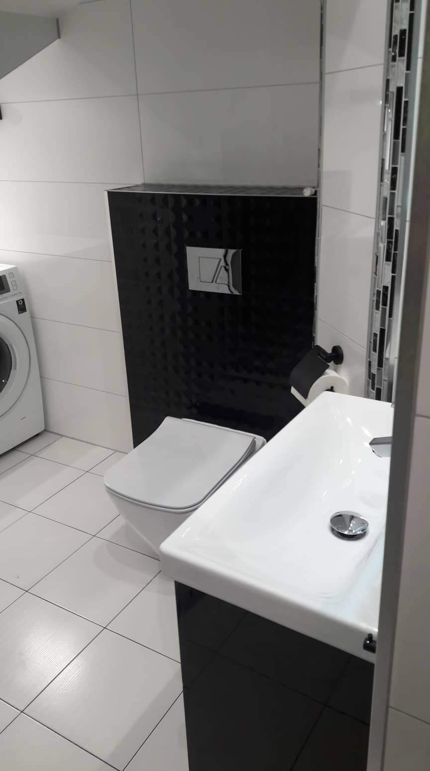 Nowoczesna łazienka z białymi płytkami, czarnym akcentem na ścianie za toaletą, białą umywalką z czarną szafką, pralką w tle i dekoracyjnym paskiem mozaiki.