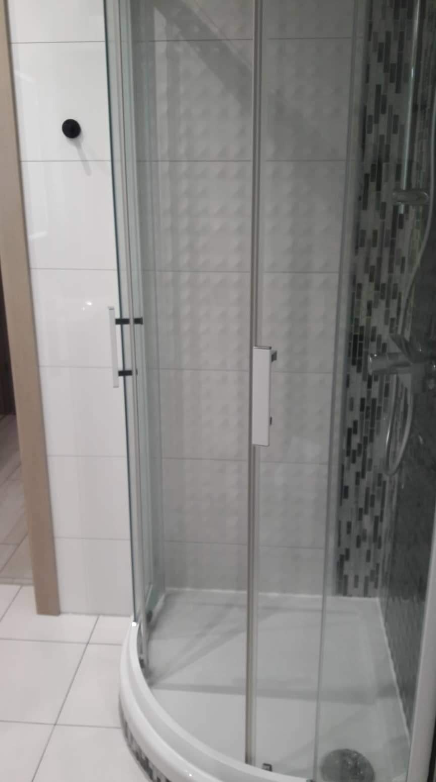 Szklana kabina prysznicowa z chromowanymi elementami, widok na białe i szare płytki z geometrycznym wzorem oraz mozaikę w odcieniach szarości i czerni.