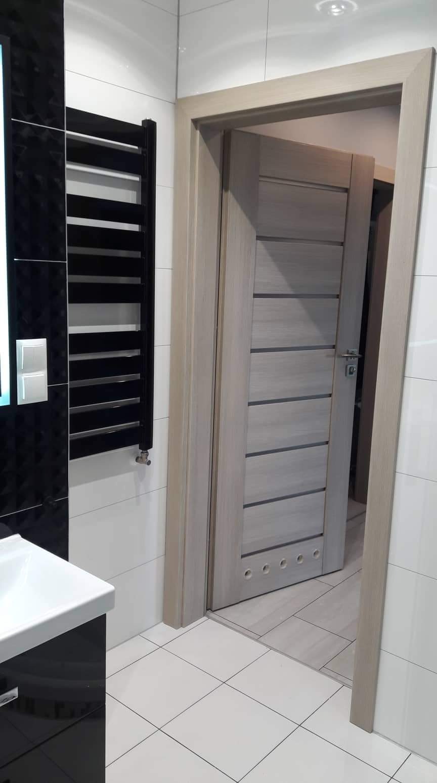 Nowoczesna łazienka z czarno-białą armaturą, grzejnikiem drabinkowym i drewnopodobnymi drzwiami wewnętrznymi z poziomymi frezami, widoczne białe płytki podłogowe i ścienne.