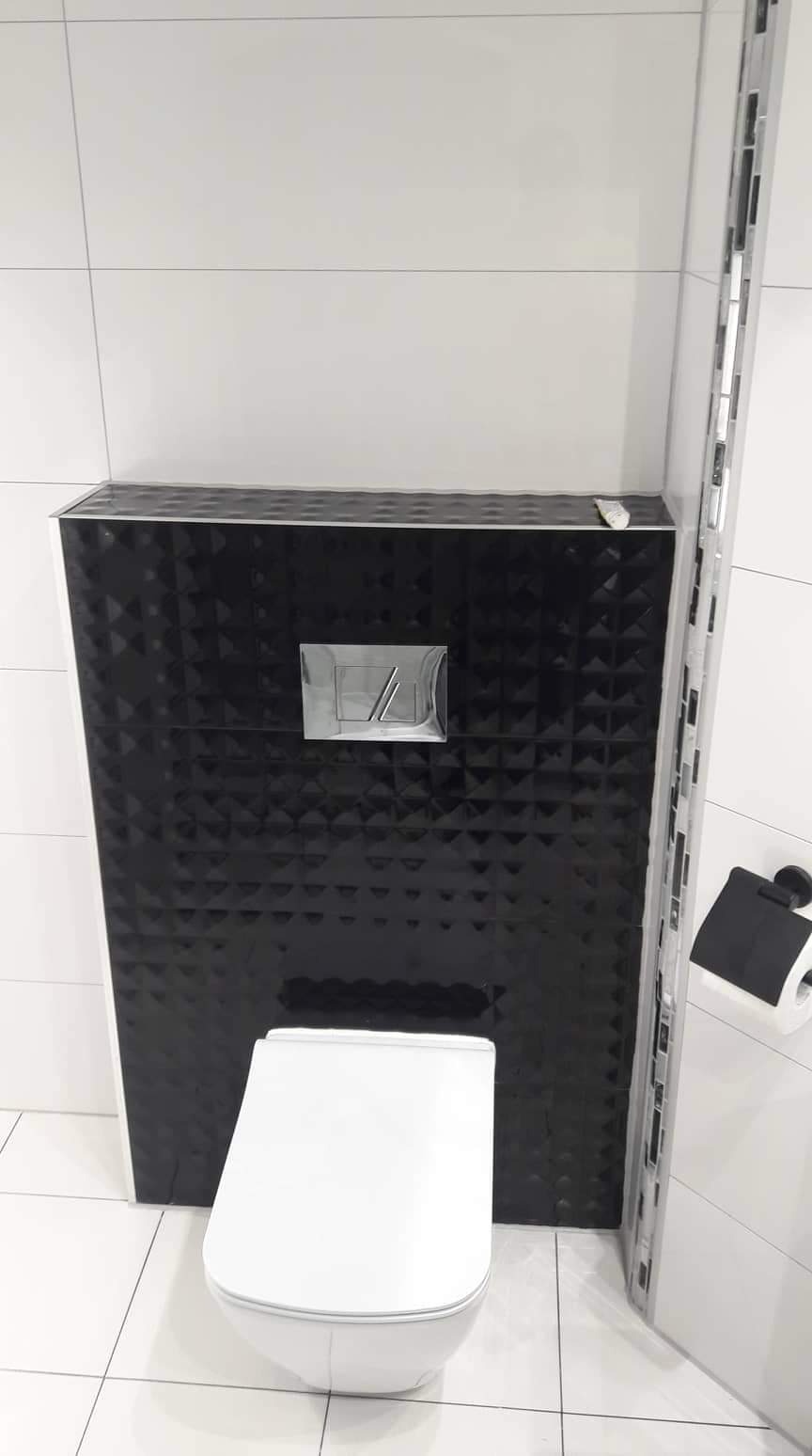 Nowoczesna toaleta podwieszana z czarną, geometryczną obudową stelaża i chromowanym przyciskiem spłukującym, białe płytki na ścianach i podłodze, dekoracyjna listwa mozaikowa z boku.