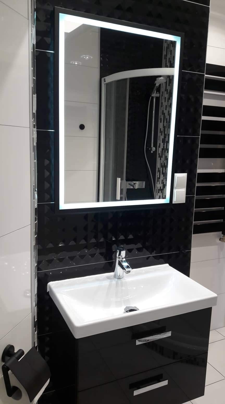 Nowoczesna łazienka z czarno-białymi płytkami, umywalką na szafce z chromowanymi uchwytami i podświetlanym lustrem, w którym odbija się kabina prysznicowa z deszczownicą.