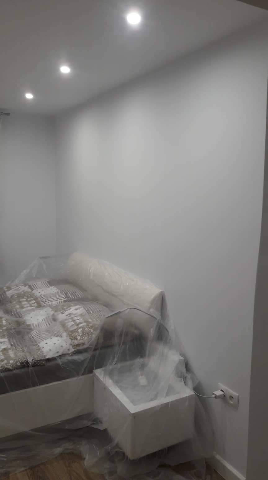 Pokój w trakcie odświeżania: ściany pomalowane na biało, meble zabezpieczone folią malarską, widoczne gniazdko elektryczne i trzy punktowe źródła światła w suficie.