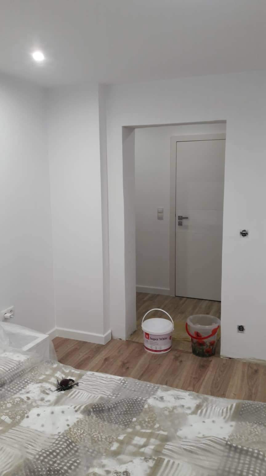 Wnętrze po świeżym malowaniu na biało, widoczne wiaderka z farbą Super White i zabezpieczona folią podłoga, widok na sąsiednie pomieszczenie z białą ścianą i drzwiami.