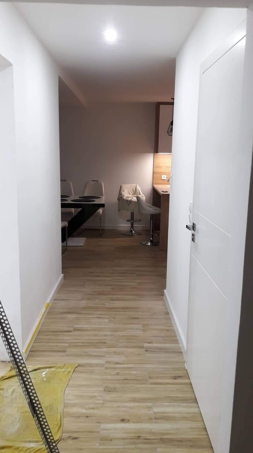 Korytarz po remoncie z nową podłogą z paneli imitujących drewno, widoczny fragment kuchni z barkiem i krzesłem barowym, oraz fragment osłony narożnika i żółta szmatka.