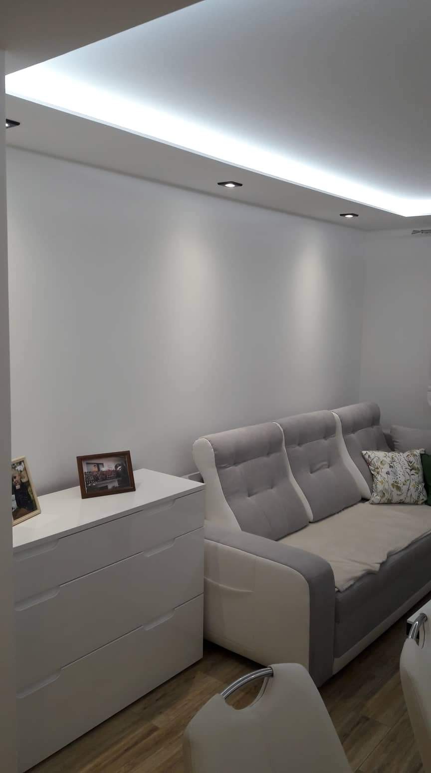 Jasne, świeżo pomalowane wnętrze pokoju w Opolu z podwieszanym sufitem z oświetleniem LED, białą komodą z ramkami i szarą sofą z poduszkami.