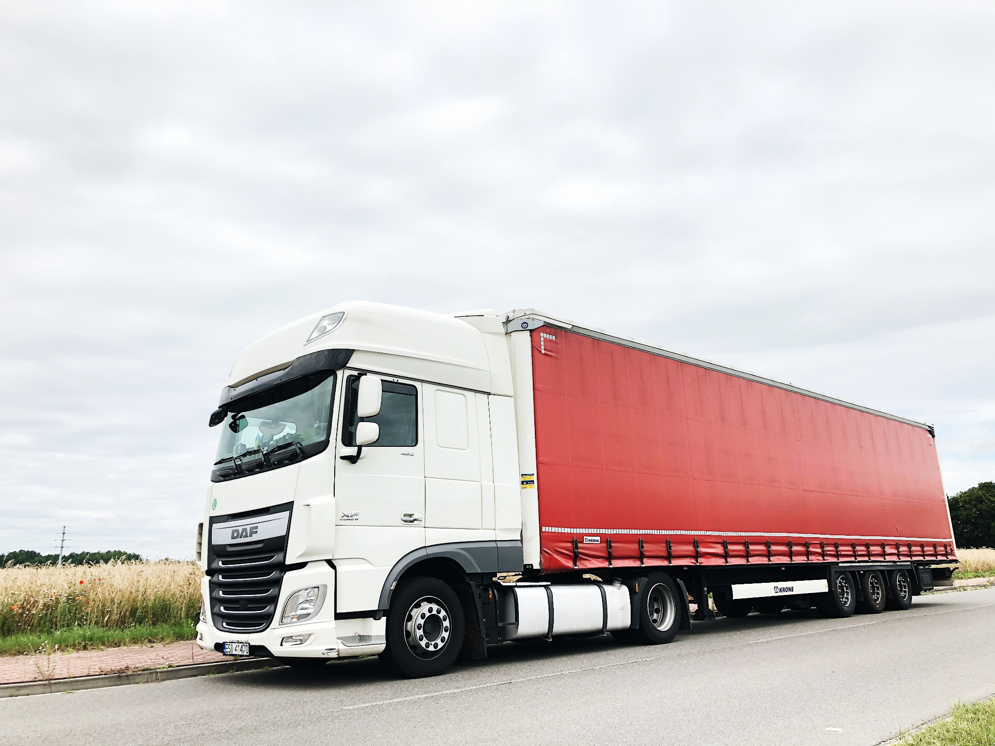 Biała ciężarówka DAF z czerwoną naczepą Krone, zaparkowana na poboczu drogi, z polem zboża w tle.