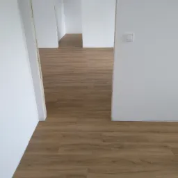 Ułożone panele podłogowe w jasnym, odnowionym pomieszczeniu z białymi ścianami i widocznymi otworami drzwiowymi prowadzącymi do kolejnych pokoi.