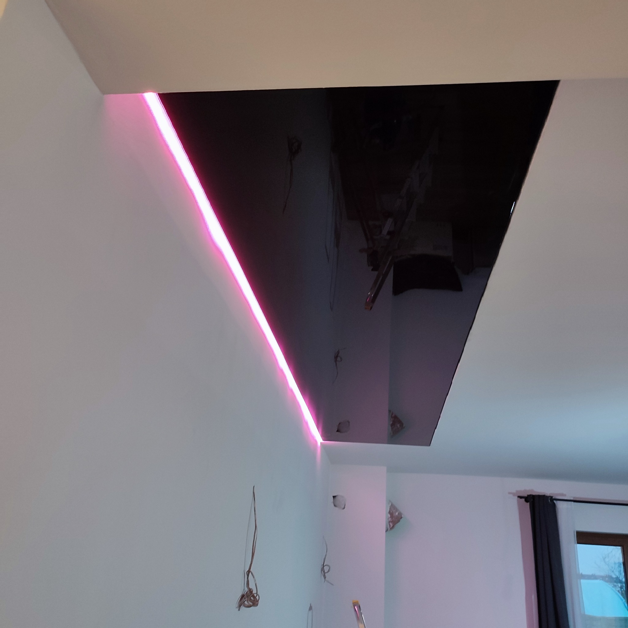 Częściowo ukończony sufit podwieszany z czarną, błyszczącą powierzchnią i wbudowanym oświetleniem LED w kolorze różowym, widoczne niedokończone instalacje elektryczne na ścianie.