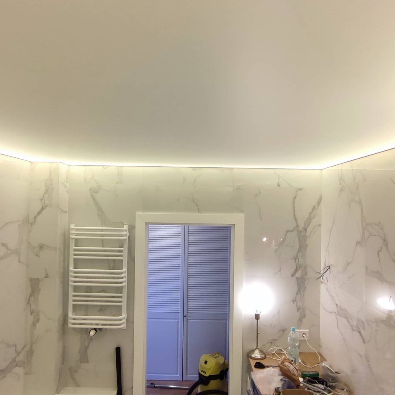 Wykończona łazienka z marmurowymi płytkami, podwieszanym sufitem z oświetleniem LED, białym grzejnikiem drabinkowym i niebieskimi drzwiami lamelowymi.