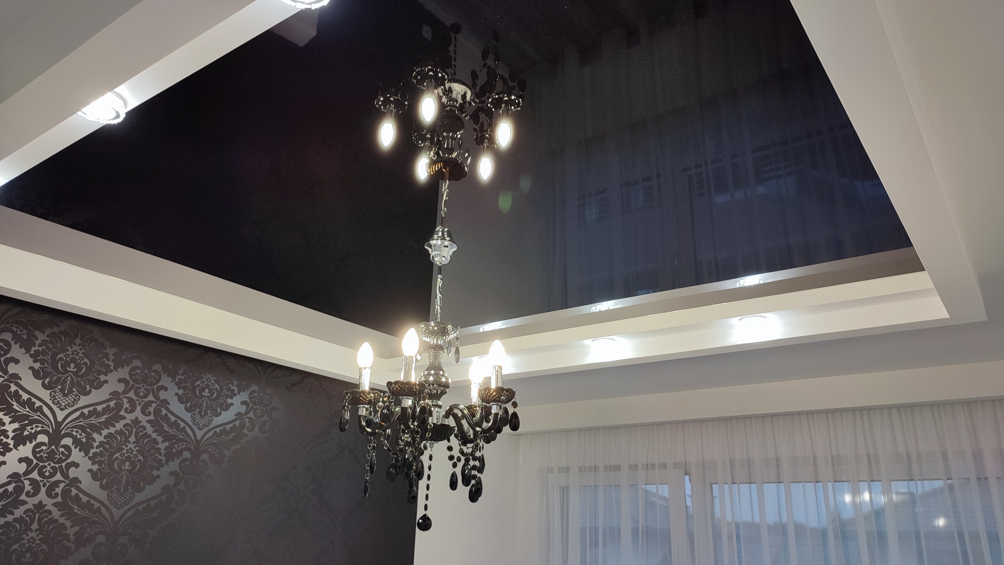 Czarny, lustrzany sufit napinany z eleganckim żyrandolem, obramowany białymi listwami i oświetleniem punktowym, w tle wzorzysta tapeta i okno z firanką.