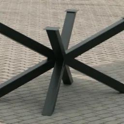 Metalowa podstawa stołu z czterema nogami w kształcie litery X, pomalowana na ciemny kolor, ustawiona na kostce brukowej.