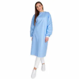 Uśmiechnięta kobieta prezentuje jasnoniebieski, jednorazowy fartuch medyczny z długimi rękawami i białymi mankietami, ubrana w białe spodnie i sportowe buty na białym tle.